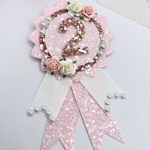 May include: Pink and white glitter ribbon with a pink glitter circle featuring a large glittery number two and a floral embellishment. The ribbon has white satin ribbon accents and pearl embellishments.