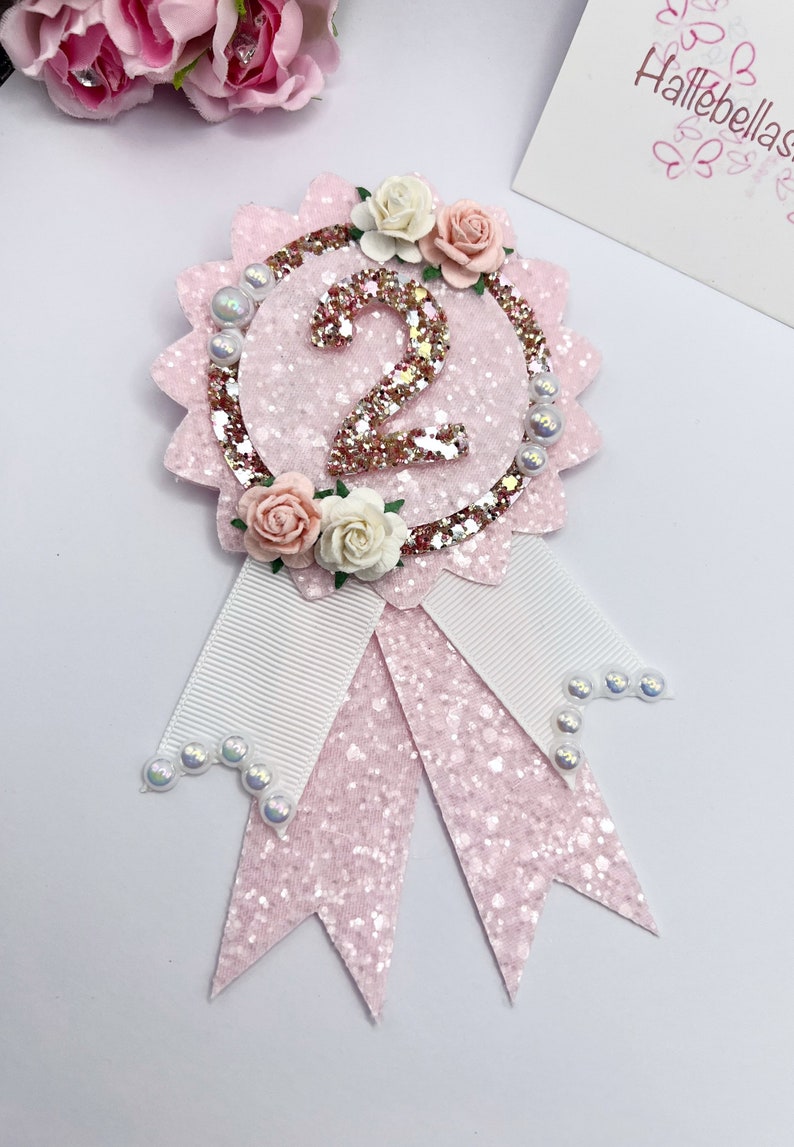 May include: Pink and white glitter ribbon with a pink glitter circle featuring a large glittery number two and a floral embellishment. The ribbon has white satin ribbon accents and pearl embellishments.