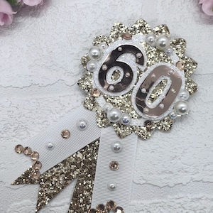 May include: Gold glitter and white ribbon rosette with the number 60 in the center, decorated with pearls and rhinestones.
