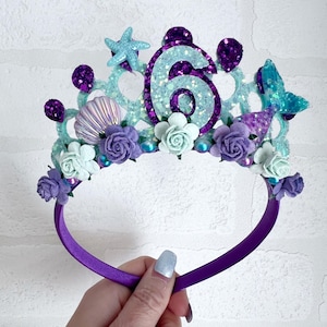 May include: Purple headband with a light blue and purple glitter crown featuring a seashell, a starfish, a mermaid tail, and the number 6.