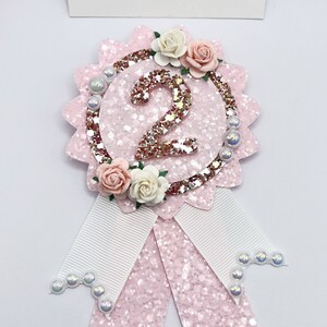 May include: Pink glitter and white ribbon rosette with a gold glitter number 2 and pink and white flowers. The rosette is decorated with white pearl beads.