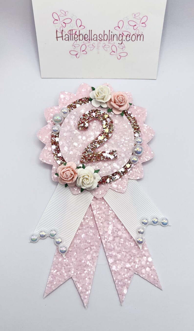 May include: Pink glitter and white ribbon rosette with a gold glitter number 2 and pink and white flowers. The rosette is decorated with white pearl beads.