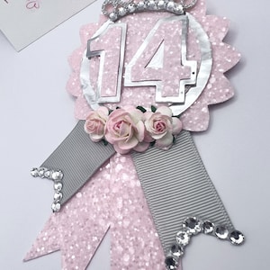 May include: Pink and silver glitter birthday badge with the number 14, a silver crown, and a gray and pink ribbon with rhinestones.