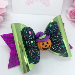 Könnte beinhalten: Eine grüne und violette Glitzer Schleife mit einer schwarzen und grünen Glitzer Mitte. Ein kleiner orangefarbener Kürbis mit einem violetten Hut und einem lächelnden Gesicht ist an der Mitte der Schleife befestigt.