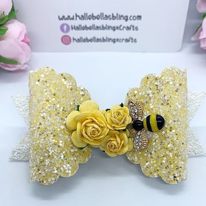 May include: A yellow glitter bow with a white lace trim and a bee charm with yellow and black stripes. The bow is decorated with yellow fabric flowers.