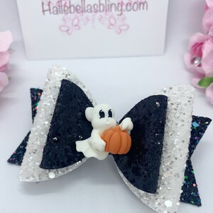 May include: Black and white glitter bow with a white ghost holding an orange pumpkin. The bow has a white glitter centre and black glitter edges. The ghost has black eyes and a small smile. The bow is perfect for Halloween.