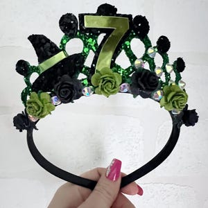 May include: Black headband with a green and black crown featuring a witch's hat, green roses, and a glittery number 7.