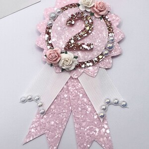 May include: Pink glitter and white ribbon rosette with a gold glitter number 2 and white pearl accents. The rosette is decorated with pink and white paper flowers.