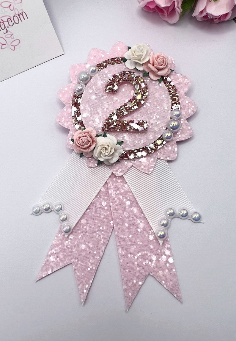 May include: Pink glitter and white ribbon rosette with a gold glitter number 2 and white pearl accents. The rosette is decorated with pink and white paper flowers.