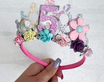 Puede incluir: Una diadema rosa con una corona plateada decorada con purpurina, flores y un gran número 5 rosa.