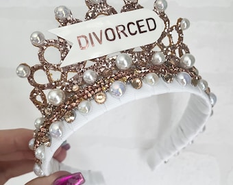Könnte beinhalten: Ein weißes Stirnband mit einer goldenen Glitzerkrone und Perlenakzenten. Die Krone hat ein weißes Banner mit dem Wort "DIVORCED" in goldenen Buchstaben.
