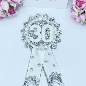 May include: Silver glitter and white satin ribbon with a white number 30 in the center. The number 30 is surrounded by white pearls.