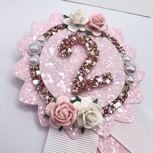 May include: Pink and white felt rosette with a glittery gold number 2 in the center. The rosette is decorated with pink and white flowers and pearl-like beads. A white ribbon is attached to the bottom.