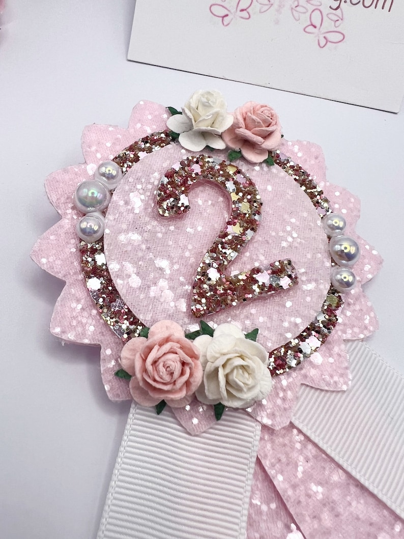 May include: Pink and white felt rosette with a glittery gold number 2 in the center. The rosette is decorated with pink and white flowers and pearl-like beads. A white ribbon is attached to the bottom.