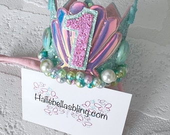Peut inclure: Une couronne de sirène rose et verte avec un numéro un pailleté sur le dessus. La couronne est décorée de perles et de coquillages irisés. La couronne est sur un bandeau rose. Le texte "hallebellasbling.com" est sur une carte blanche sous la couronne.