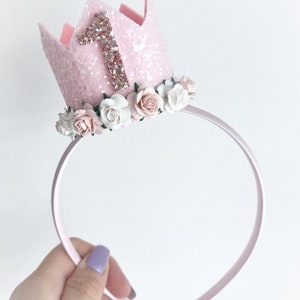 May include: Pink felt crown with a glitter finish and a pink felt number one on top. The crown is decorated with a circle of white and pink felt flowers and is attached to a pink headband.