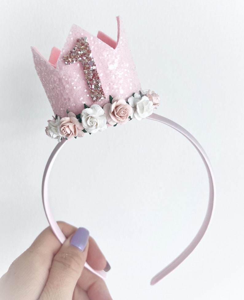 May include: Pink felt crown with a glitter finish and a pink felt number one on top. The crown is decorated with a circle of white and pink felt flowers and is attached to a pink headband.