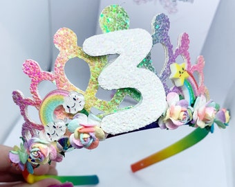 Puede incluir: Diadema de arcoíris con un número 3 blanco con purpurina, flores y un arcoíris con una nube. La diadema está decorada con purpurina y tiene un diseño de arcoíris.