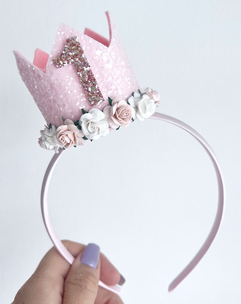 May include: Pink felt crown headband with a glittery number one and a floral trim. The headband is made of a thin, flexible plastic.