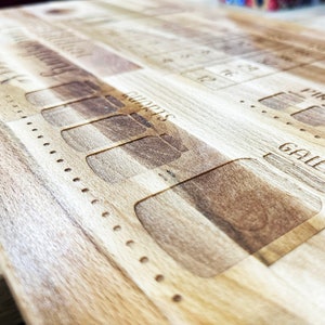 May include: Wooden cutting board with engraved markings for liquid measurements, including quarts, pints, and gallons. The board has a light brown color and a natural wood grain pattern. The design also includes a series of numbered markings.