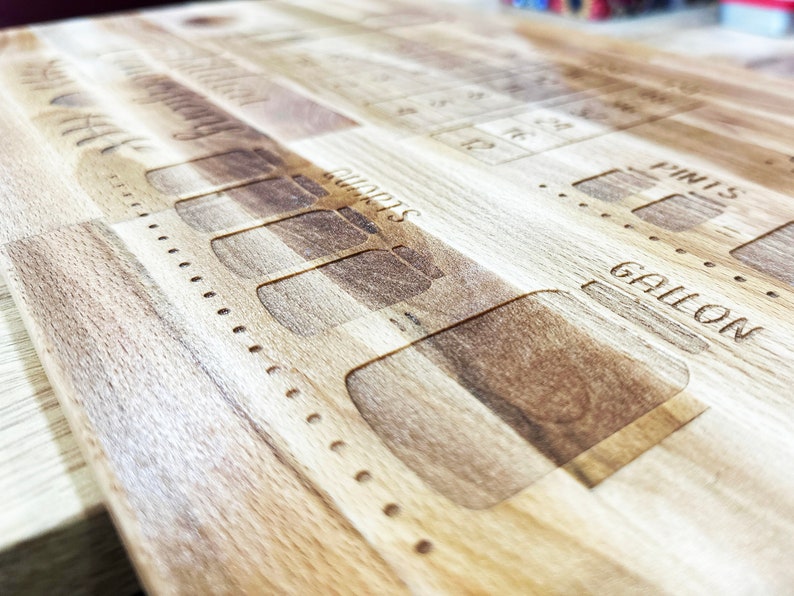 May include: Wooden cutting board with engraved markings for liquid measurements, including quarts, pints, and gallons. The board has a light brown color and a natural wood grain pattern. The design also includes a series of numbered markings.