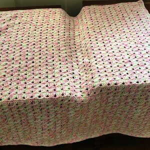 May include: A crocheted baby blanket in pink, white and green. The blanket is made with a delicate stitch pattern and has a soft, fluffy texture.