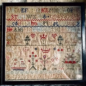 以下が含まれることがあります： 黒いフレームに入ったアンティークのサンプラー。赤、青、ベージュの糸でクロスステッチ刺繍が施されています。デザインには、文字、数字、木、ハート、船などのモチーフが含まれています。サンプラーは石の表面に表示されています。