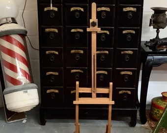 May include: A black wooden cabinet with 18 drawers, each with a gold label. A wooden easel stands in front of the cabinet. A red and white striped barber pole is on the left side of the image.