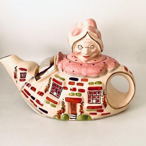 Può includere: Una teiera in ceramica a forma di casa con un viso di donna sul tetto. La teiera è decorata con mattoni rossi, erba verde e finestre bianche. La donna ha le guance rosa e un cappello rosa.