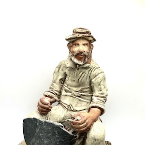 May include: A ceramic figurine of a man sitting on a rock, wearing a hat and holding a small object in his hand. The figurine is painted in shades of brown, white, and gray.