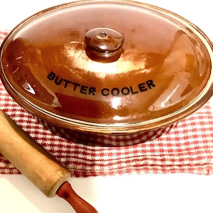 Può includere: Un refrigeratore per burro in ceramica marrone con un coperchio e le parole "Butter Cooler" stampate sul coperchio. Il refrigeratore è appoggiato su un panno a quadri rosso e bianco accanto a un mattarello di legno.