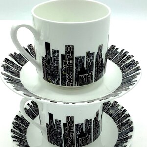 Könnte beinhalten: Ein Set aus zwei weißen Keramik-Teetassen und Untertassen mit einem schwarz-weißen Stadtbild-Design. Die Tassen haben einen Henkel und die Untertassen einen gewellten Rand.