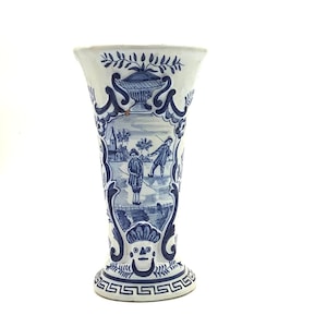 May include: A white ceramic vase with a blue and white floral design. The vase has a flared top and a round base. The design features a scene of two figures in a landscape.