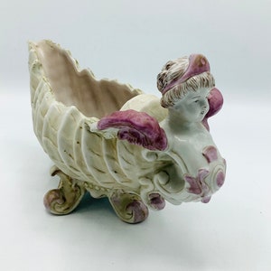 May include: A porcelain figurine of a woman in a shell-shaped planter. The woman is wearing a pink and white dress and has a crown on her head. The planter is decorated with pink and white flowers.