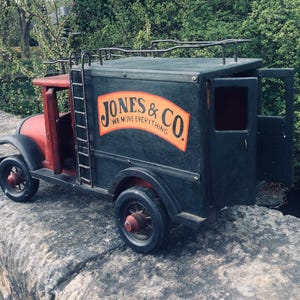 May include: A vintage wooden toy truck, painted in red and black, with the text "JONES & CO WE MOVE EVERYTHING" on the side. The truck has a ladder, open door, and a roof rack. The toy is parked on a stone surface.