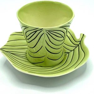 Puede incluir: Un juego de taza y platillo de té verde con un diseño de hoja. La taza y el platillo están decorados con líneas negras que crean un patrón de hoja.