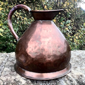 May include: An antique copper pitcher with a bulbous body and a flared spout. It has a curved handle and a circular base. The copper has a reddish-brown patina. The pitcher is likely of European origin. The background shows green foliage and branches.