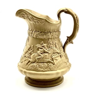 Puede incluir: Una jarra de cerámica de color crema con un diseño en relieve detallado de un caballo y un jinete. La jarra tiene un pico ancho y acampanado y una asa decorativa.