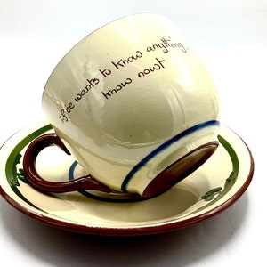 Puede incluir: Una taza de té y un platillo de cerámica blanca con un asa marrón. La taza de té tiene una franja azul y blanca alrededor del borde y el texto "If ee wants to know anything, know nowt" escrito en marrón en el lateral.