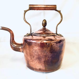May include: A copper kettle with a curved spout and a handle. The kettle has a lid with a small knob on top. The handle is made of a lighter-coloured metal. The kettle is likely vintage or antique.