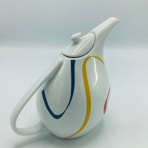 May include: White ceramic teapot with a curved spout and handle. The teapot features a colourful abstract design with blue, yellow, and red lines.