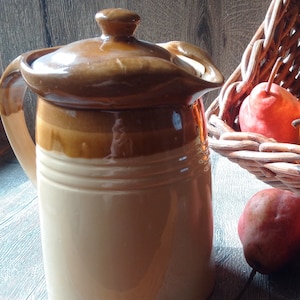 May include: A brown and beige ceramic pitcher with a lid. The pitcher is sitting on a wooden surface next to a wicker basket filled with pears.