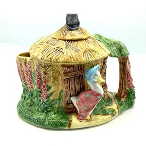 Può includere: Una teiera in ceramica a forma di cottage con il tetto di paglia con un'oca che indossa un cappello e un vestito rosa in piedi davanti alla porta. La teiera ha una base verde e una porta marrone.