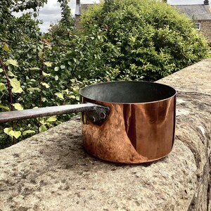 May include: A copper pot with a long wooden handle. The pot has a shiny, slightly tarnished finish and is sitting on a stone surface.