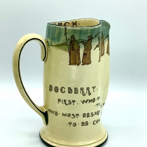 Może przedstawiać: Dzbanek ceramiczny w kolorze kremowym z brązowym uchwytem i zielono-niebieską glazurą. Dzbanek ma cytat po boku, który brzmi "DOGBERRY FIRST. WHO. THE MOST DESAR. TO. BE. COM."