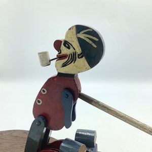 May include: A wooden toy figure of a man with a red shirt, blue pants, and a black hat. He is holding a pipe in his mouth and is sitting on a wooden base. The figure is painted in bright colors and has a whimsical expression.