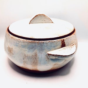 Könnte beinhalten: Weiße Keramik-Auflaufform mit Deckel und zwei Griffen. Die Form hat ein rustikales, handgeworfenes Aussehen mit braunen Glasur-Akzenten.