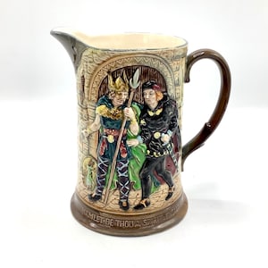 May include: A ceramic jug with a brown handle and a scene from Hamlet. The scene depicts two men in medieval clothing, one holding a spear. The text "HAMLET-BE THOU A SPIRIT SPEAK" is on the bottom of the jug.