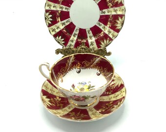 May include: A red and gold teacup and saucer set with a floral design. The saucer has a scalloped edge and a red and gold striped pattern. The teacup has a floral design and a gold rim.