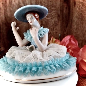 May include: A porcelain figurine of a woman wearing a blue hat and a white and blue dress with lace trim. The figurine is sitting on a white base.
